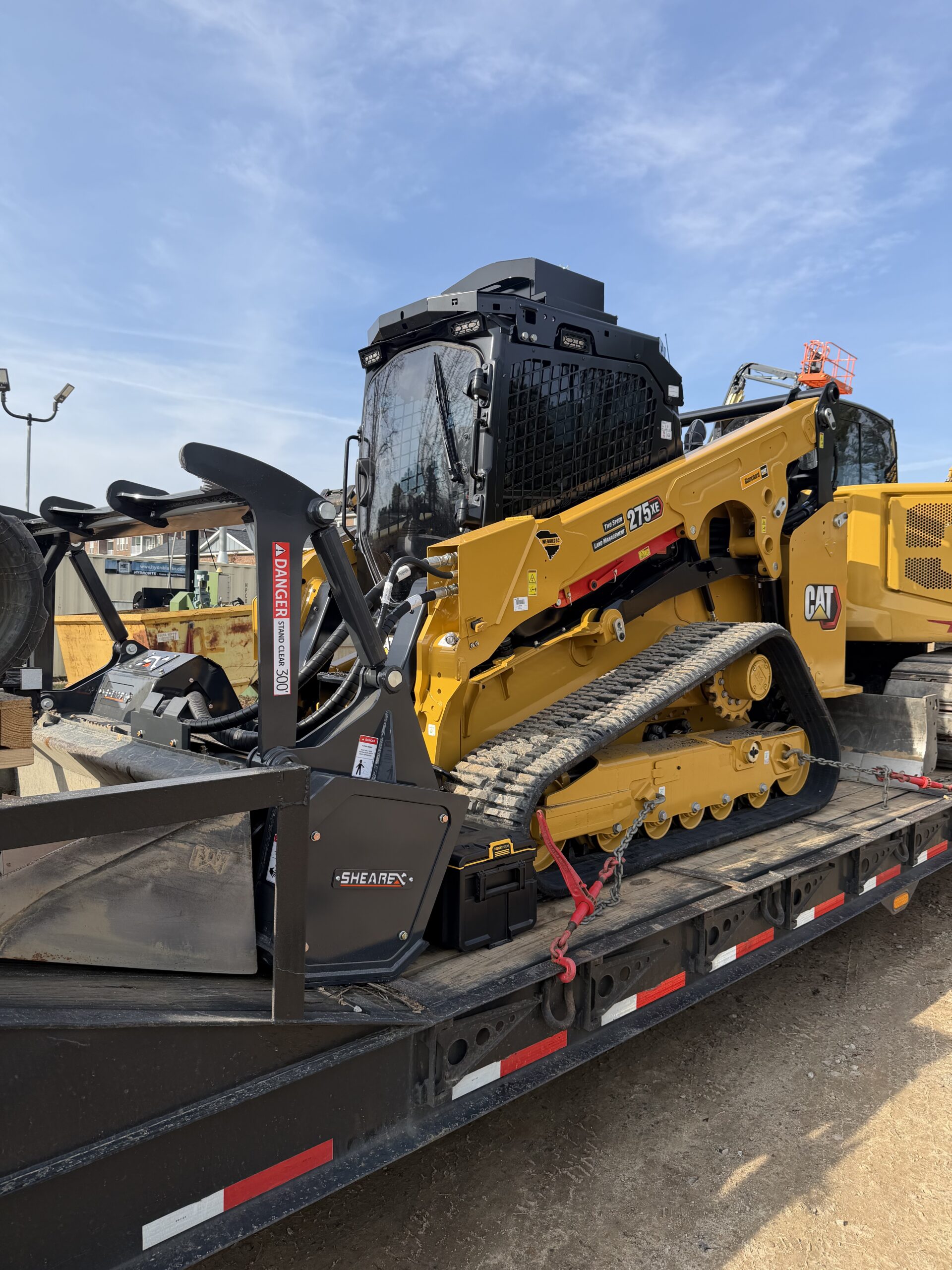 A yellow Caterpillar 279D3 compact track loader is secured on a flatbed trailer at a construction site, with other equipment visible in the background.