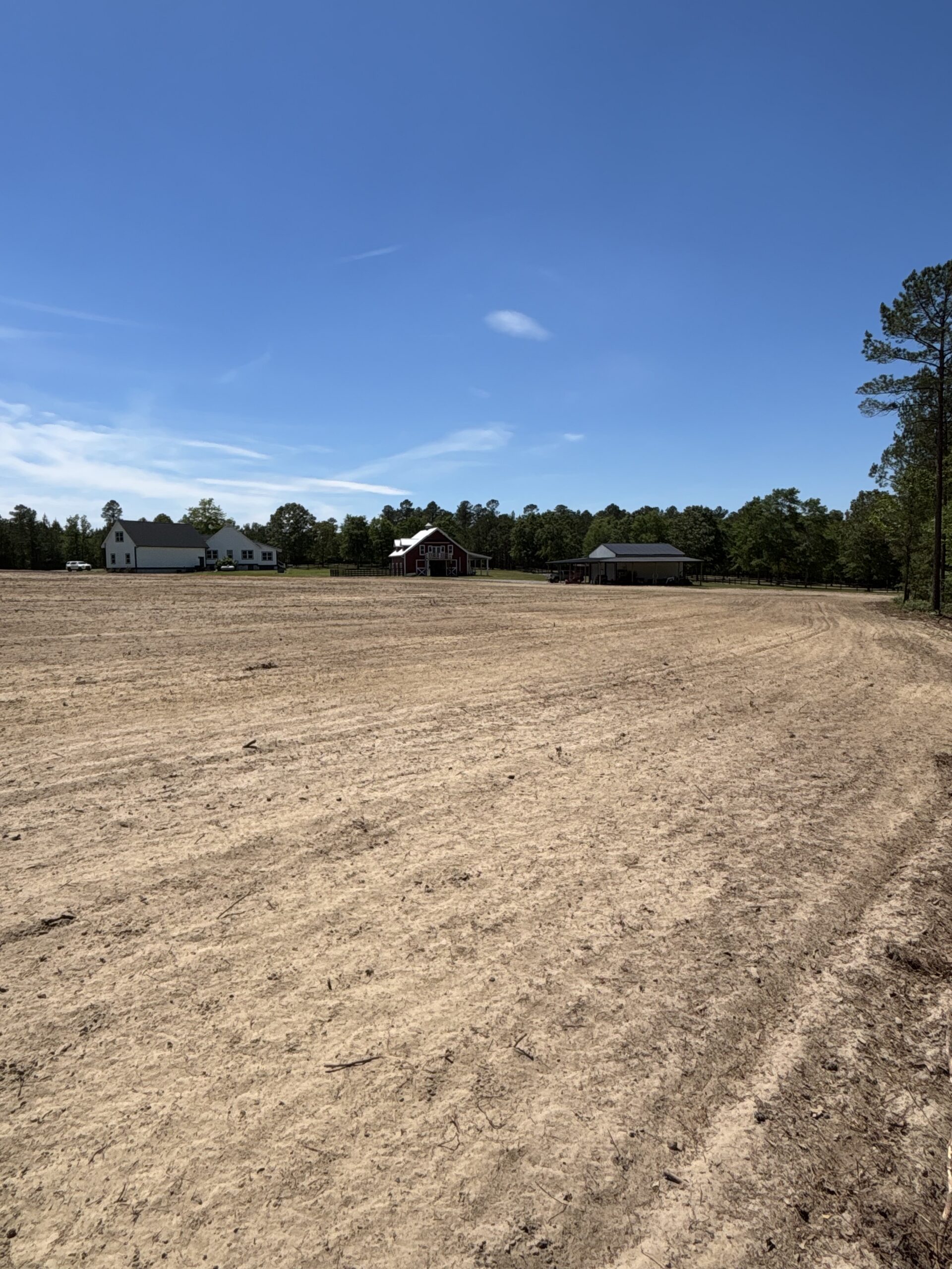 A large, dry, plowed field under a clear blue sky with three houses and trees in the background.