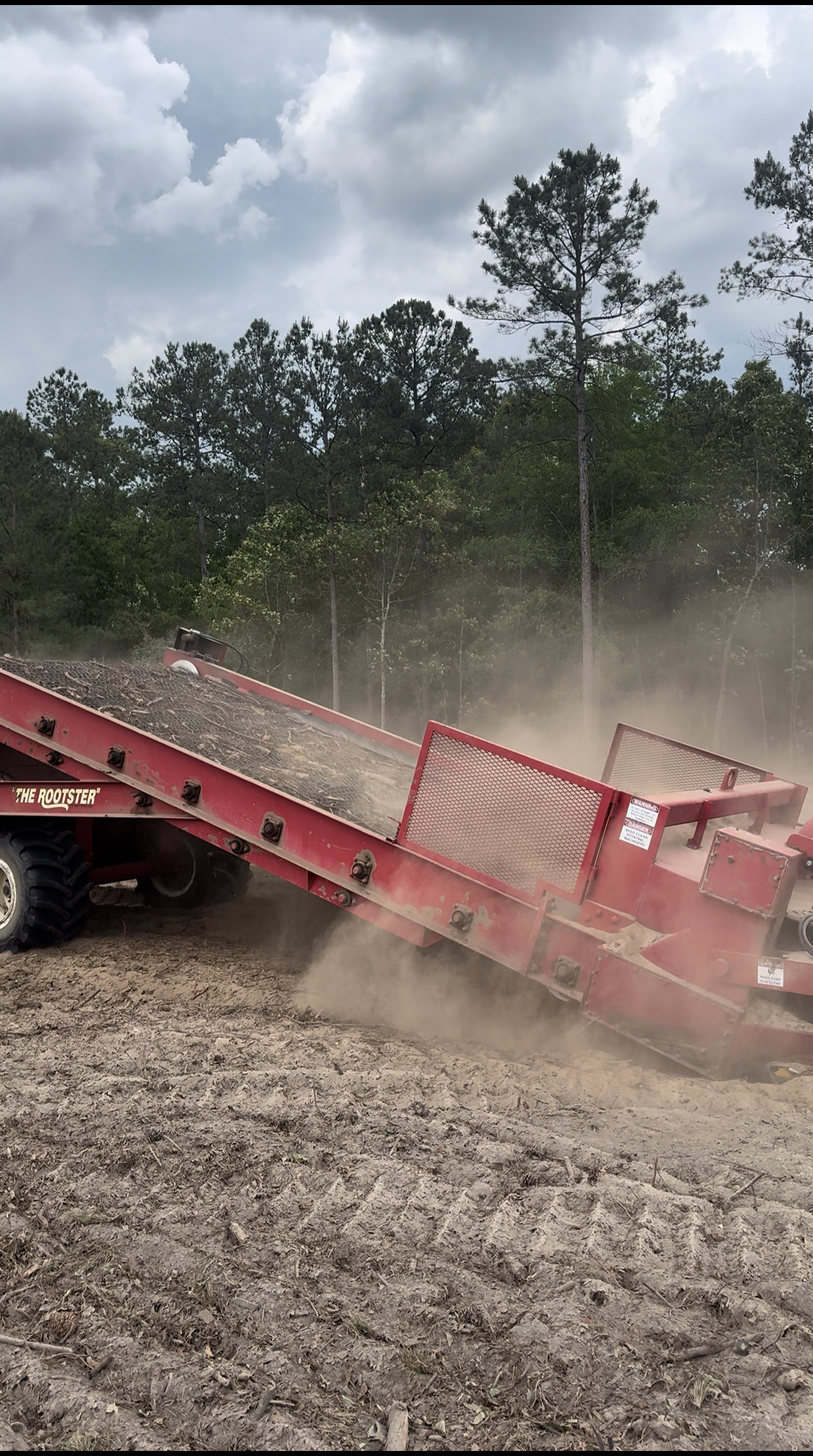A red agricultural machine spreads soil on a dusty field, with trees and a cloudy sky in the background.